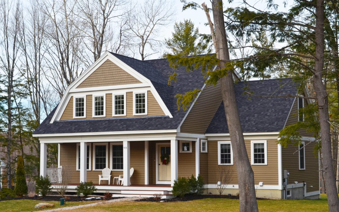 A picturesque home on a wooded lot with light brown siding and contrasting white windows, a wide front porch and dark gray roof.