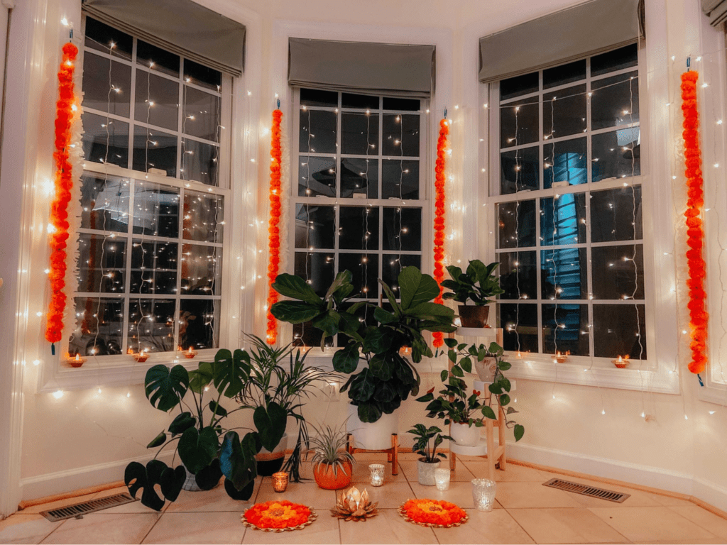 There are twinkle lights and orange Diwali decor hanging across three windows. There are also green plants set on the floor in front of the windows, and candles on the window sills
