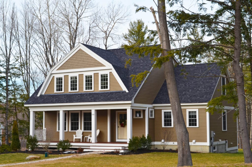 A picturesque home on a wooded lot with light brown siding and contrasting white windows, a wide front porch and dark gray roof.