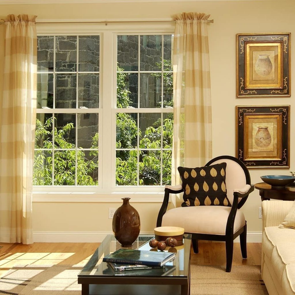 Living room with two large traditional styled windows, two hanging pictures, striped curtains and black and gold pillow laying on a white and black chair. 