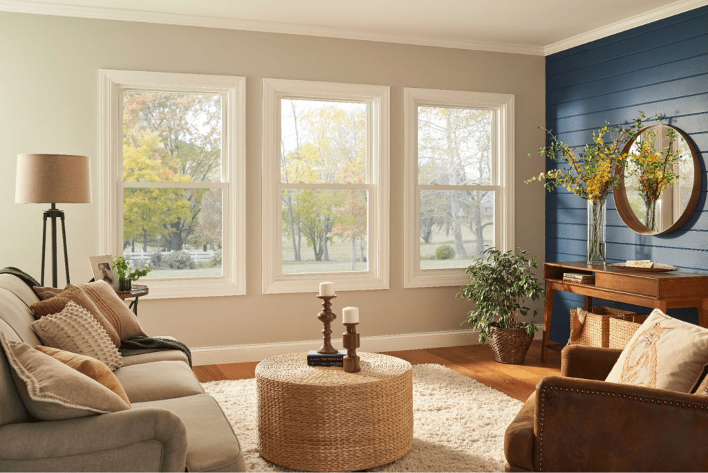 An interior view of a living room with three vinyl windows showing the fall trees outside, and a navy blue shiplap adjoining wall. There is a beige couch and armchair, and a brown desk
