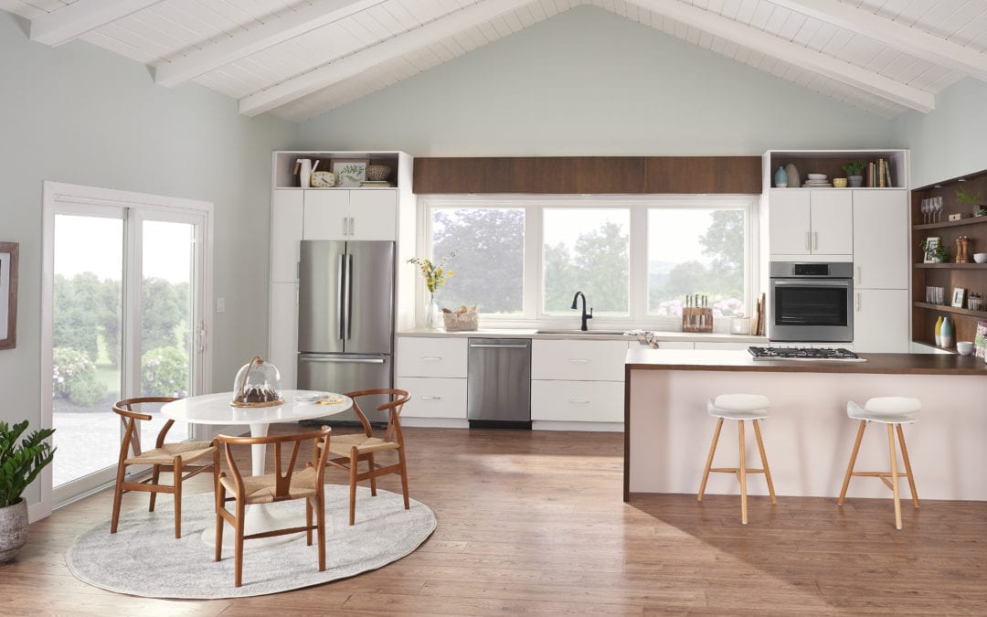 An image of a galley kitchen, showcasing stainless steel appliances, a counter with two stools, a small kitchen table to the left, a sliding glass door and multiple windows over the sink.