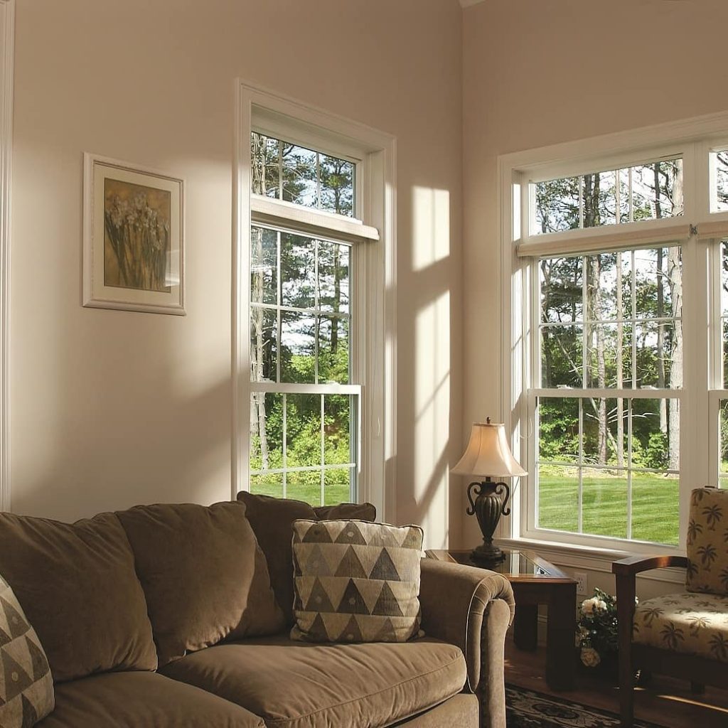 Alt text: A living room with a brown sofa, patterned pillows, a hanging picture, a side table, a lamp and another chair. There are three double-hung vinyl windows, with a transom window above each of them.
