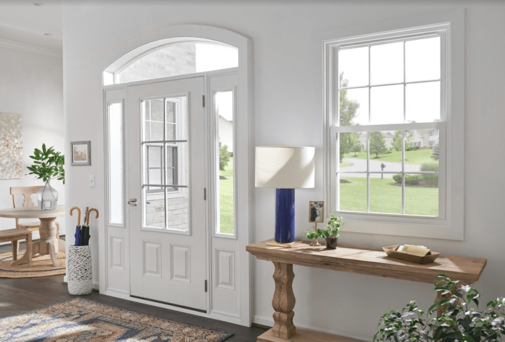 View of a white front door and entry windows from inside the home surrounded by wood flooring, colorful rug and greenery