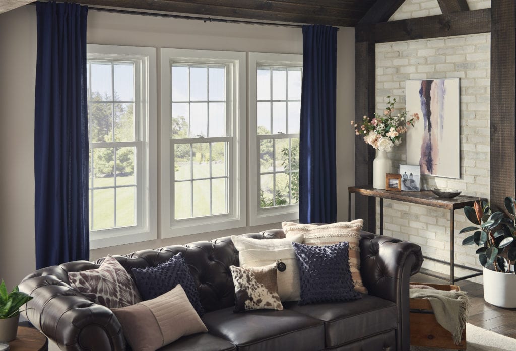 A well-designed living room with three large windows, dark blue curtains and a homey leather couch with large, inviting accent pillows.