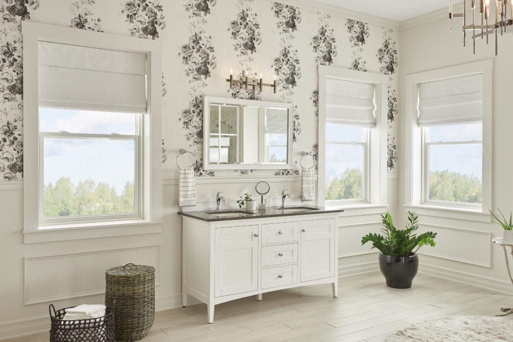 A large shabby chic bathroom with floral patterned wallpaper, a vintage-inspired bathroom cabinet and large windows overlooking the forest. 