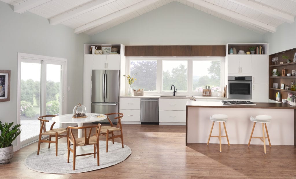An image of a galley kitchen, showcasing stainless steel appliances, a counter with two stools, a small kitchen table to the left, a sliding glass door and multiple windows over the sink.